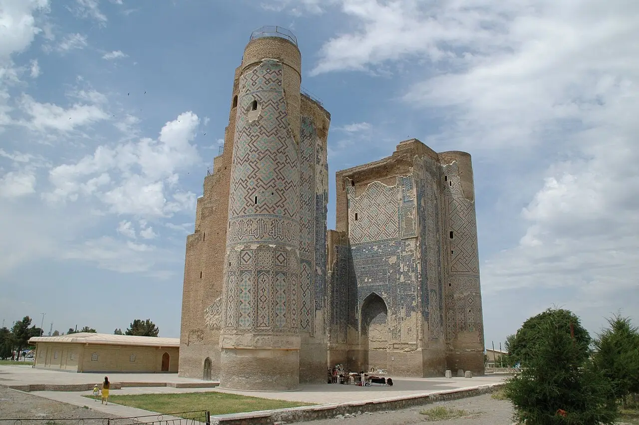 As duas torres revestidas de azulejos geométricos azuis do portal em ruínas do Ak-Saray, em Shakhrisabz, com nuvens e um pavilhão baixo ao lado.