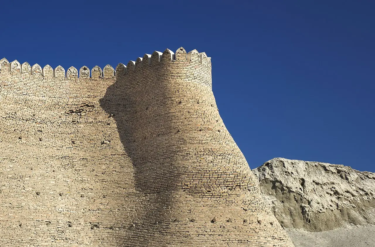 Muralha e torre redonda de tijolo com ameias da cidadela Ark de Bukhara, contra um céu azul intenso, com um monte de terra à direita.