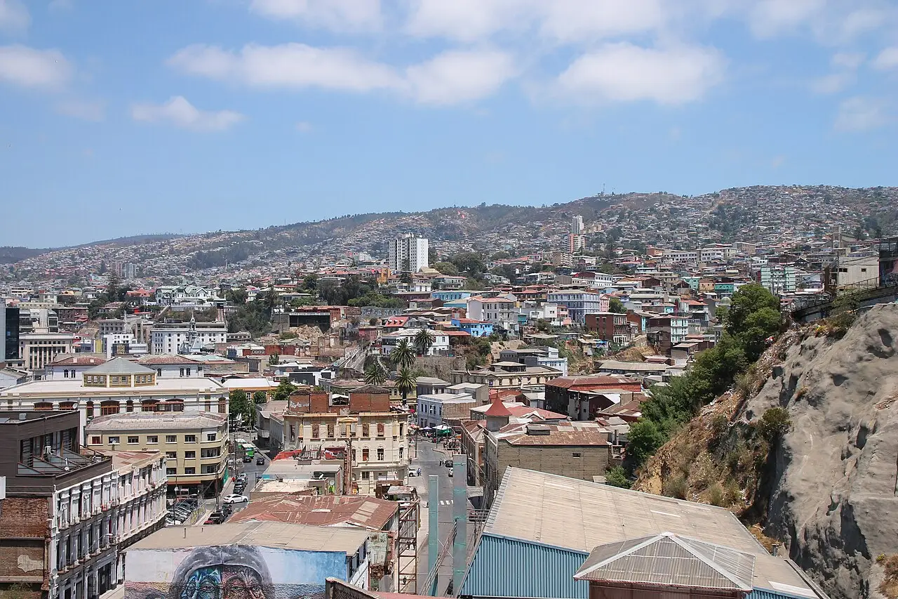 Vista de Valparaíso a partir de um mirante, com casas coloridas cobrindo os morros, prédios antigos em primeiro plano e parte de um mural na parte de baixo.