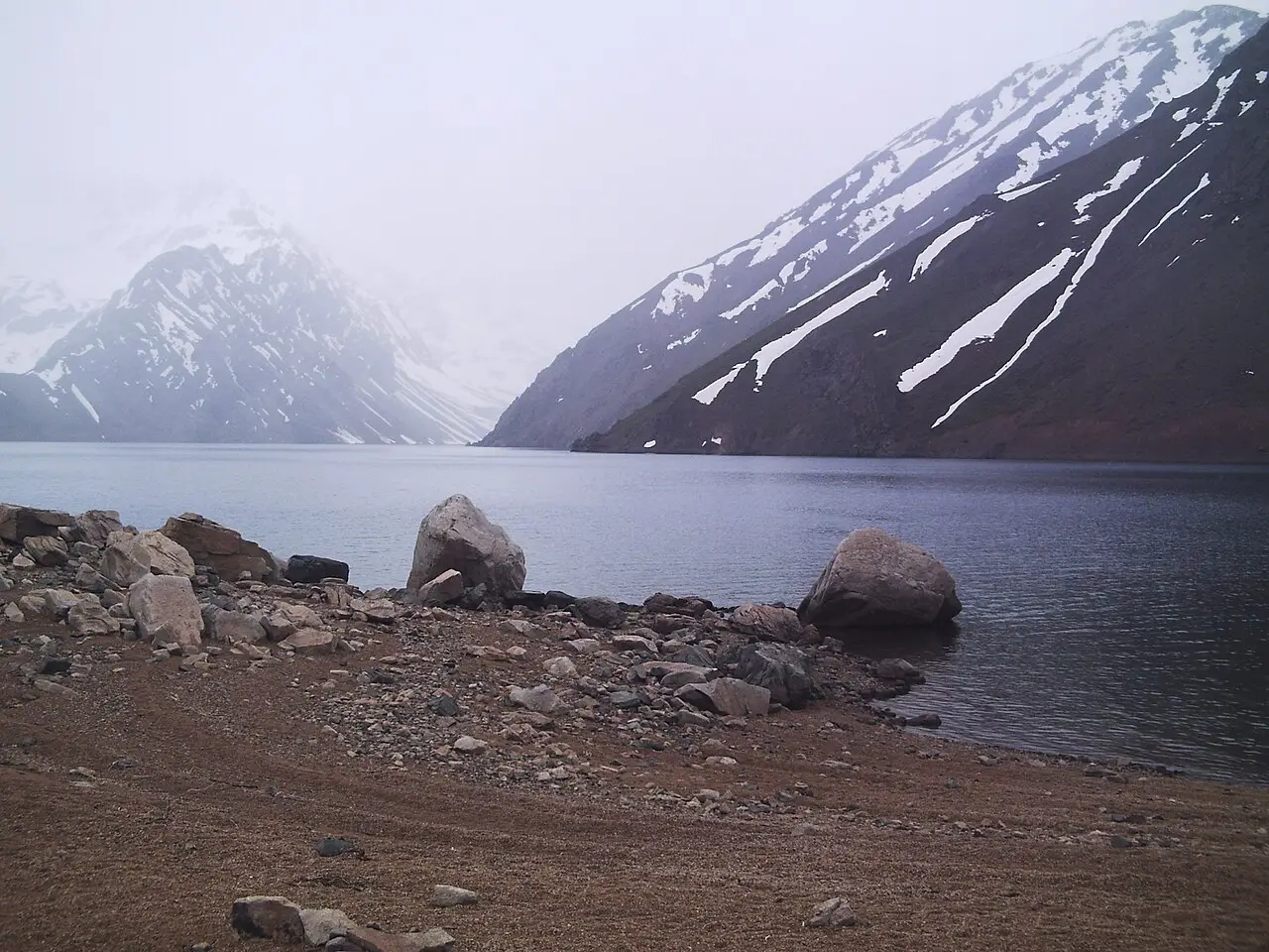 Lago de água escura entre montanhas com faixas de neve nas encostas, sob névoa, com pedras grandes e cascalho na margem, na Laguna Negra do Cajón del Maipo.