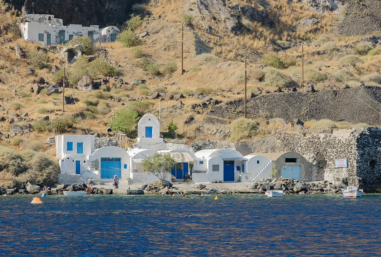Casinhas brancas com portas azuis à beira da água em Thirasia, ao pé de uma encosta seca de cascalho vulcânico, com barcos pequenos e o mar azul-escuro na frente.