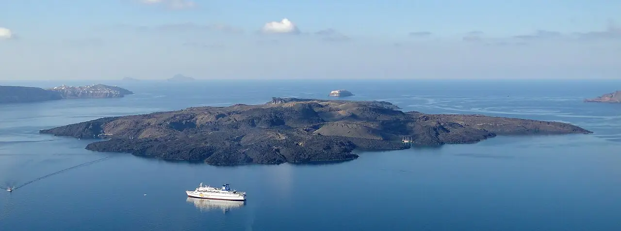 A ilha escura e plana de Nea Kameni no meio da caldeira, com um ferry branco no mar calmo e outras ilhas ao fundo.