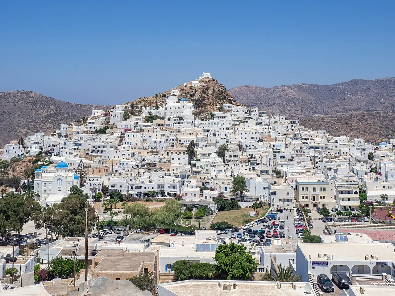 Vista da Chora de Ios, uma cidade de casas brancas cúbicas subindo por uma colina com igrejas de cúpula azul, com estacionamento em primeiro plano e serras ao fundo.