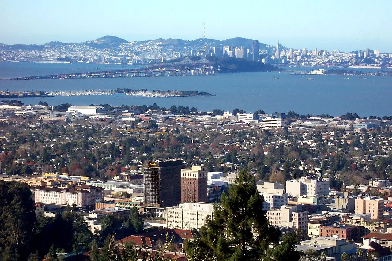 Vista do centro de Berkeley a partir das colinas, com prédios e árvores em primeiro plano, a baía e a Bay Bridge no meio e o horizonte de São Francisco ao fundo.