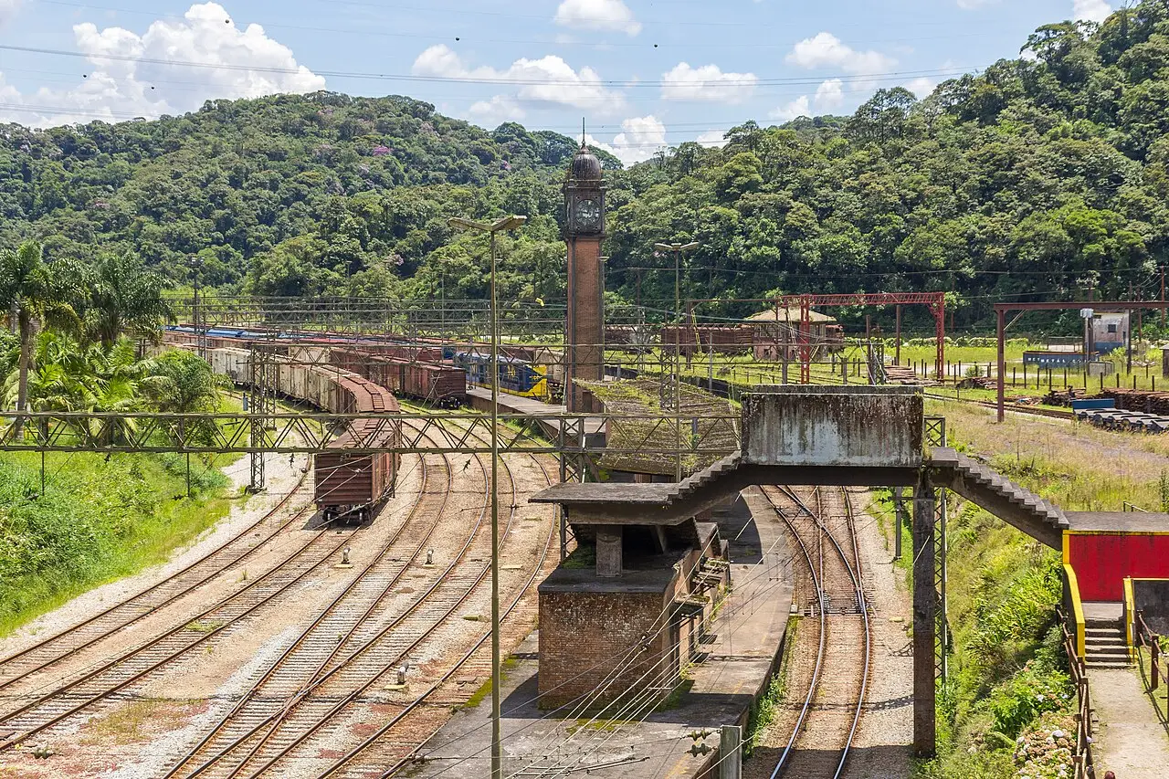 Pátio ferroviário de Paranapiacaba, com vários trilhos, vagões parados, uma passarela de concreto e a torre do relógio de tijolo diante de morros cobertos de mata.