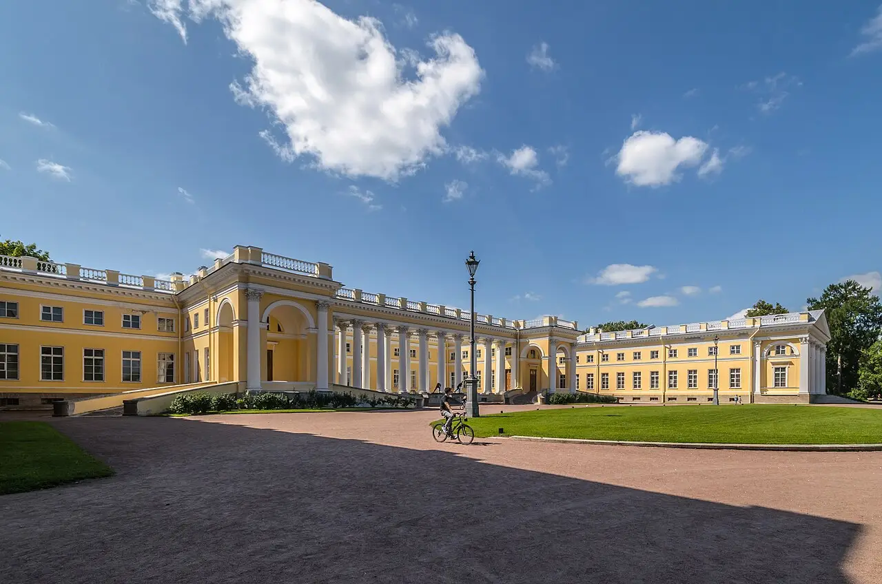 O Palácio de Alexandre, em Tsárskoie Seló, de fachada amarela com colunata branca, um gramado à direita e uma pessoa de bicicleta no caminho de cascalho.