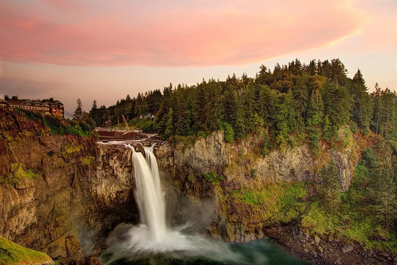 Snoqualmie Falls tombe le long d’une paroi rocheuse couverte de mousse, avec une forêt de conifères au sommet, un bâtiment dans le coin et un ciel rosé au crépuscule.