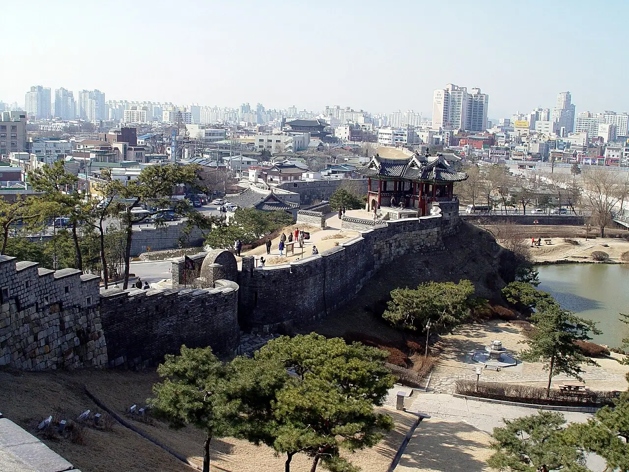 Trecho da fortaleza Hwaseong com muralha de pedra e ameias, um pavilhão de telhado curvo sobre o bastião, um lago à direita e a cidade moderna de Suwon ao fundo.