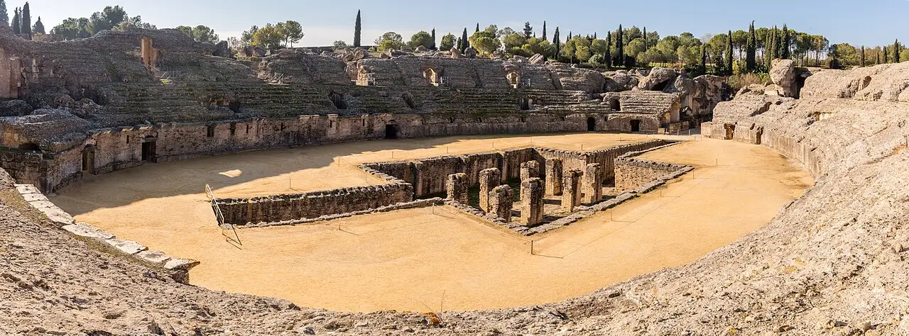 Panorama do anfiteatro romano de Itálica, com a arena de terra clara, fundações de pedra no centro, arquibancadas gastas ao redor e ciprestes no alto.