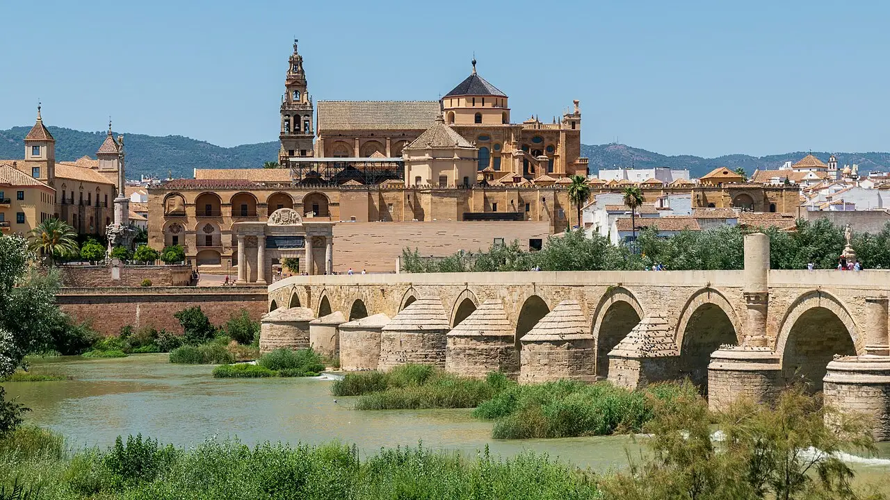 A Mesquita-Catedral de Córdoba e a ponte romana de arcos sobre o rio Guadalquivir, com o campanário e telhados de telha ao fundo sob céu azul.