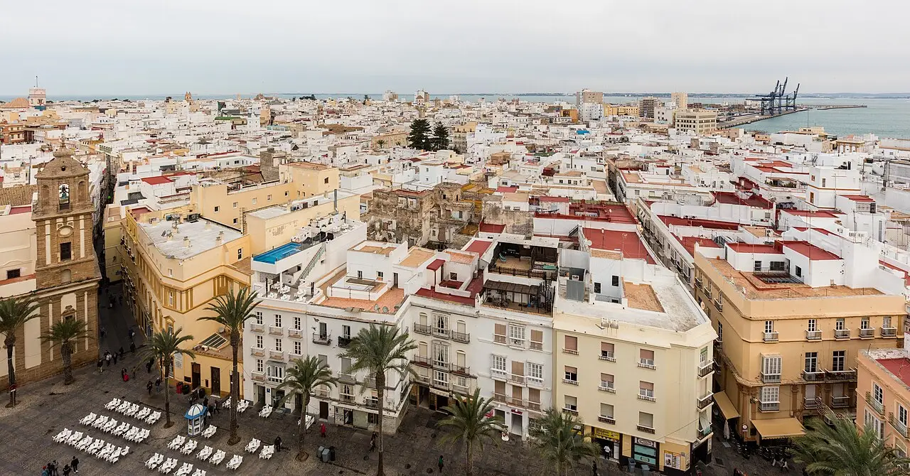 Vista aérea de Cádiz, com casas brancas e ocres de terraço plano, palmeiras e uma praça com cadeiras brancas, e o mar e um guindaste do porto ao fundo.