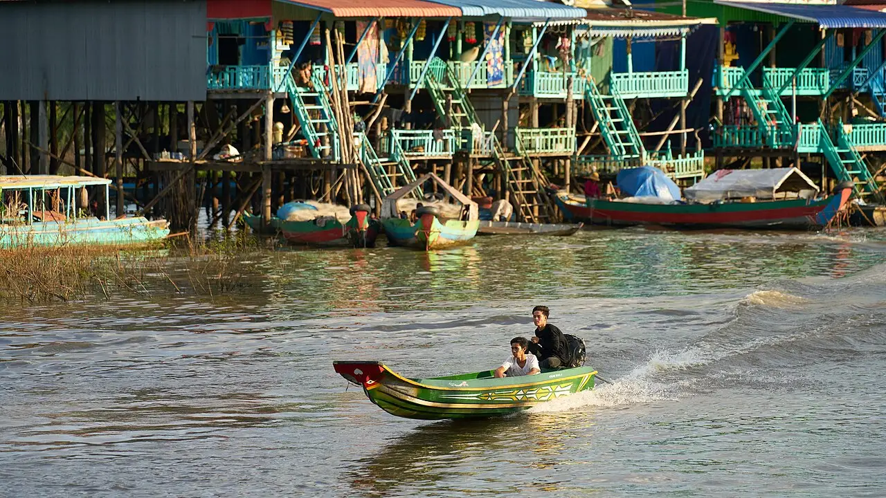 Casas de palafita coloridas, com escadas verdes, às margens de um rio marrom, com barcos amarrados e um barco a motor com dois jovens passando na frente, em Kampong Khleang.
