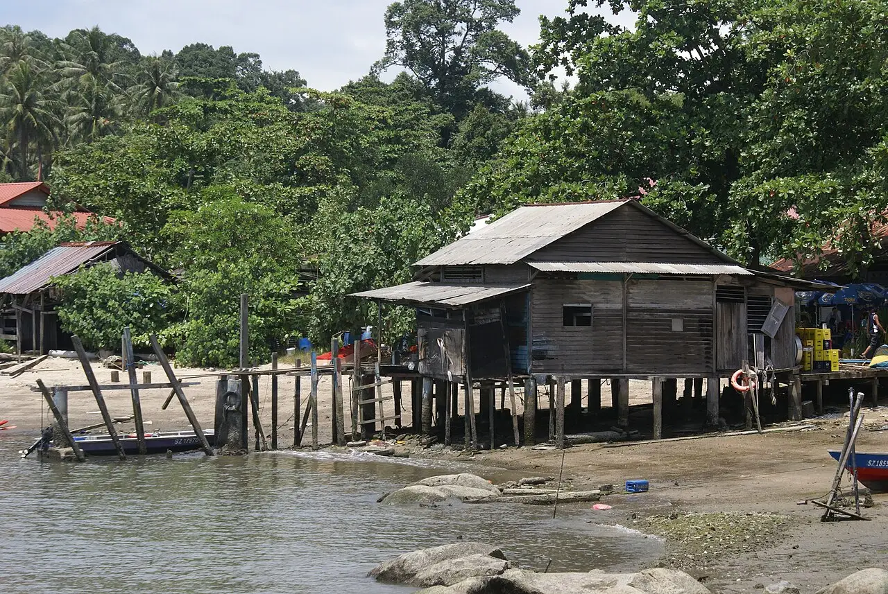 Casa de madeira sobre palafitas na beira da praia de Pulau Ubin, com telhado de zinco, um píer simples, barcos pequenos e mata densa atrás.