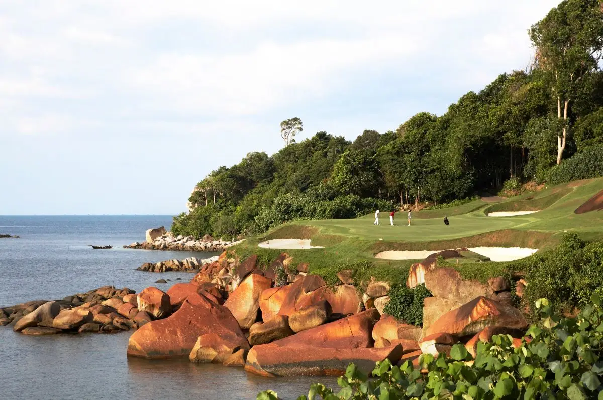 Rochas vermelhas na beira do mar e o gramado com árvores no alto da encosta.