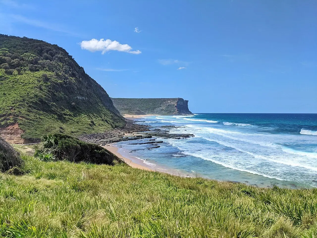 Praia estreita entre um morro coberto de mata e o mar com ondas, com plataforma de rocha na água e um promontório ao fundo, na Garie Beach, no Royal National Park.