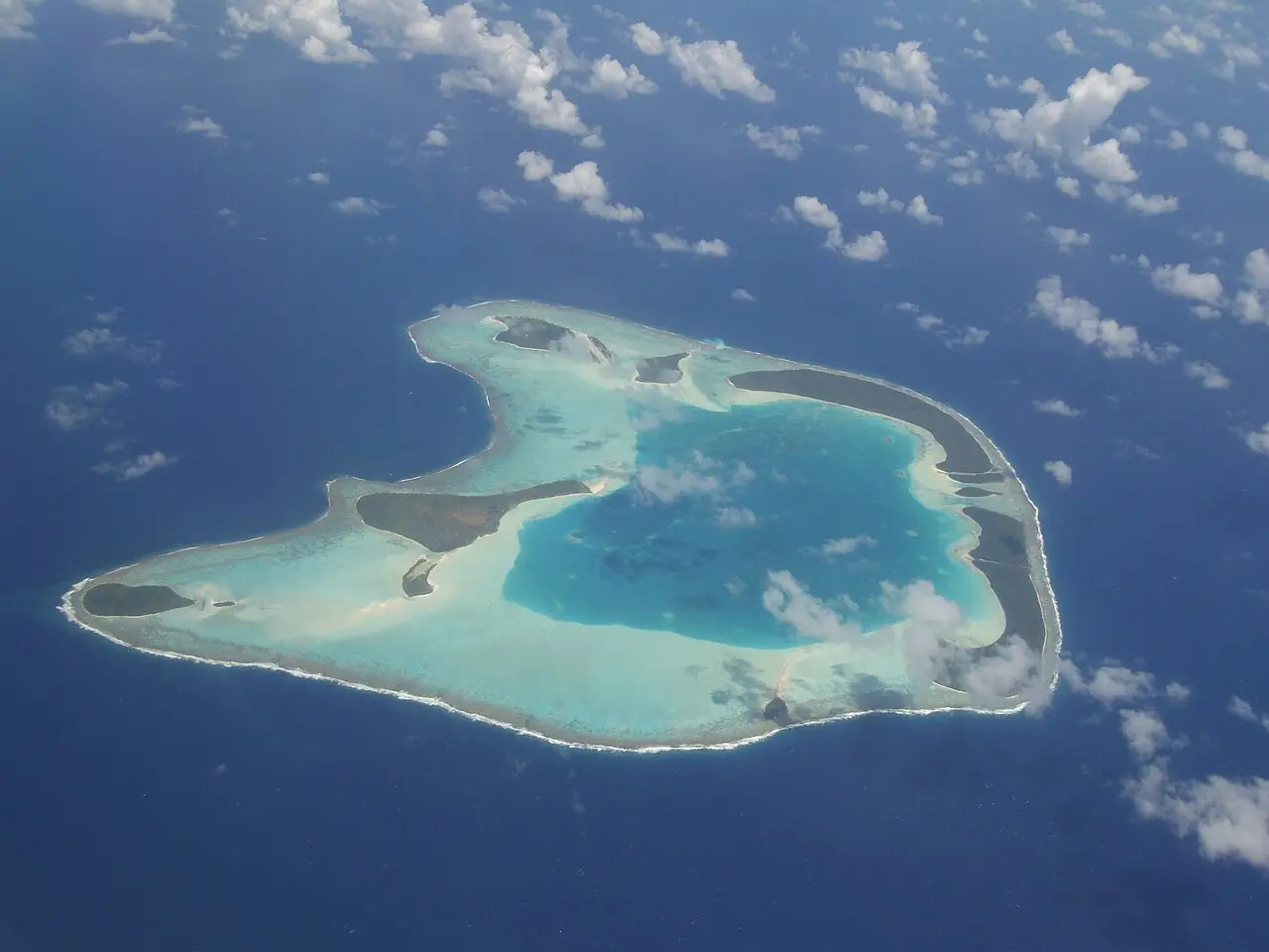 Vista aérea do atol de Tetiaroa, com um anel de recifes e ilhotas cobertas de vegetação em volta de uma laguna azul-turquesa, no oceano azul-escuro.