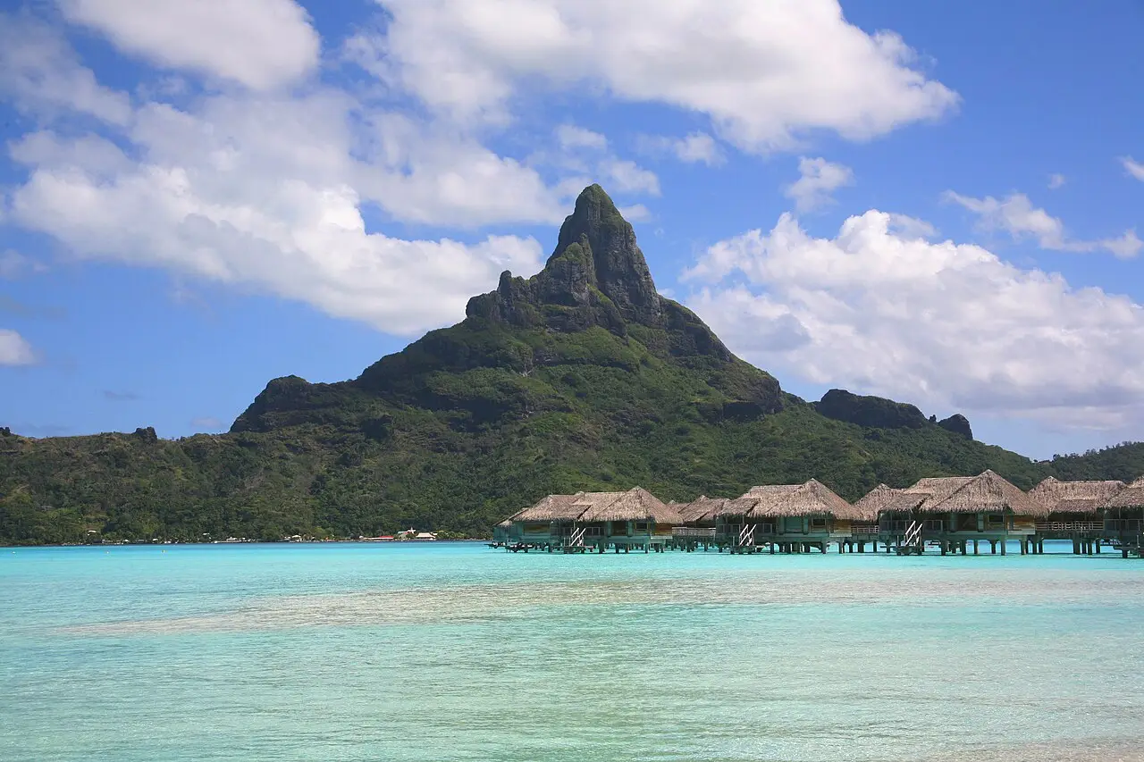 A ilha de Bora Bora vista da água, com o pico rochoso coberto de mata ao fundo, bangalôs de palha sobre a laguna e água turquesa em primeiro plano.