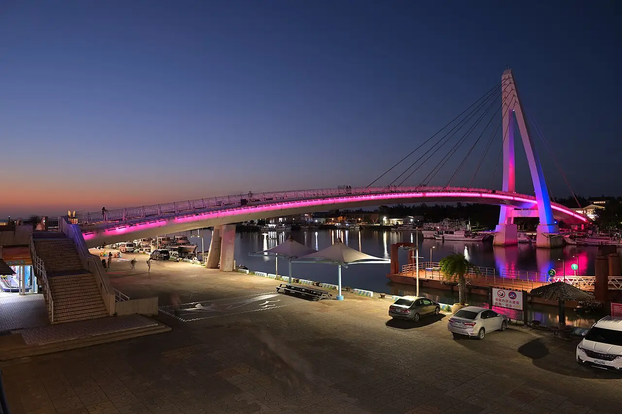 A Ponte dos Namorados de Tamsui ao anoitecer, com o arco iluminado em rosa, o mastro com cabos, o porto com barcos e alguns carros no cais.