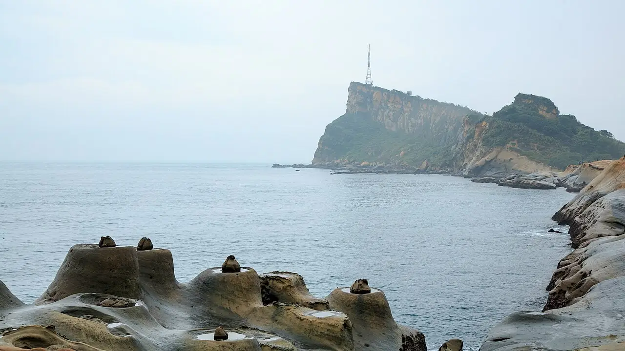 Formações de arenito em forma de cogumelo e de panela, com pequenas pedras no topo, em primeiro plano, e um promontório com torre de antena sobre o mar, em Yehliu.
