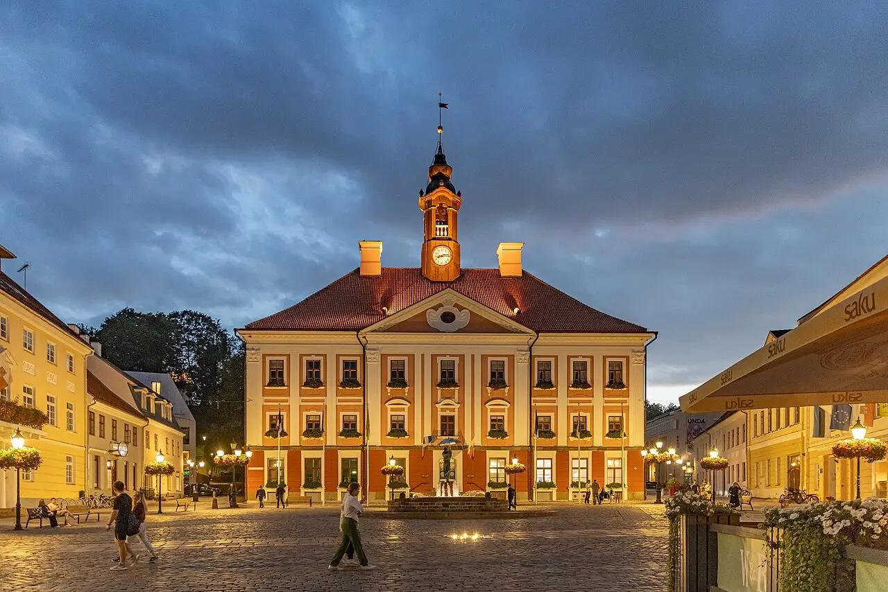 A prefeitura de Tartu ao anoitecer, edifício de fachada clara e telhado vermelho com torre de relógio, numa praça de paralelepípedos com pessoas caminhando e luzes acesas.