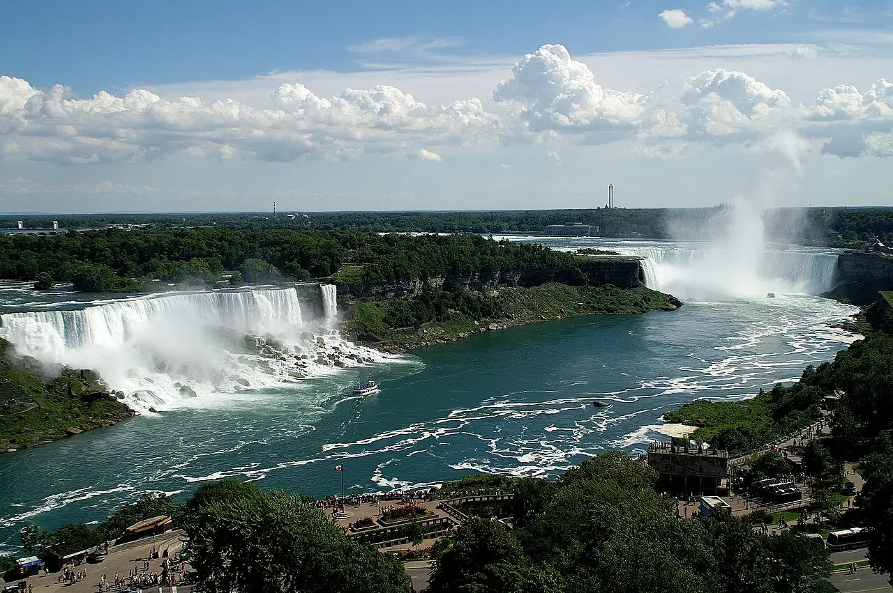 Vista desde lo alto de las cataratas del Niágara, con las caídas Americana y Velo de Novia a la izquierda, la Herradura a la derecha, un barco en el río y miradores con gente en las orillas.