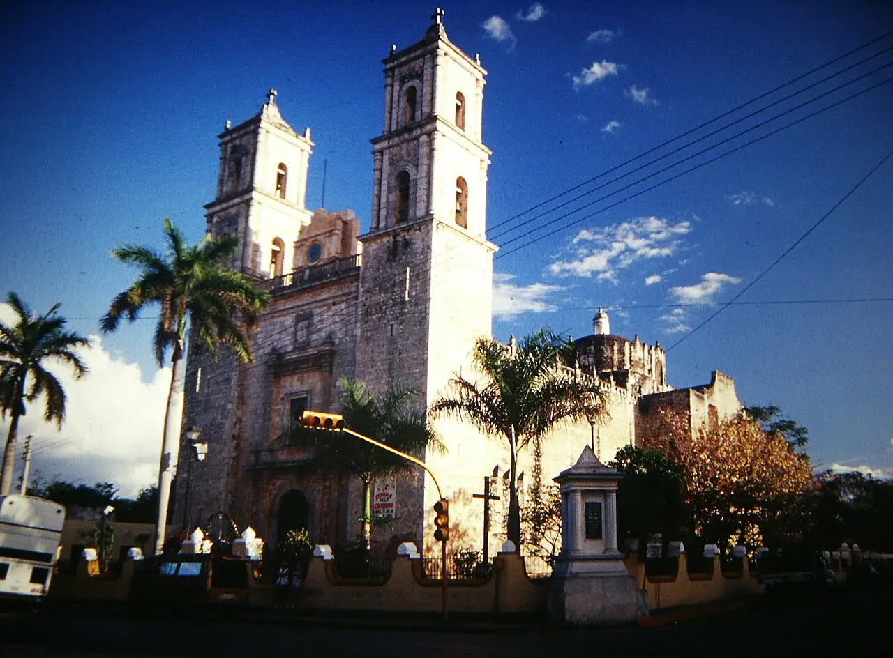 A igreja de São Servácio em Valladolid, com duas torres brancas e pedra escura, palmeiras em frente e um semáforo na esquina, sob céu azul intenso.