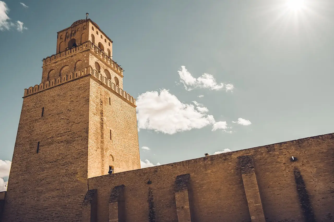 El minarete cuadrado de ladrillo de la Gran Mezquita de Kairuán y un tramo del muro, vistos desde abajo, con el sol brillando en la esquina del cielo.