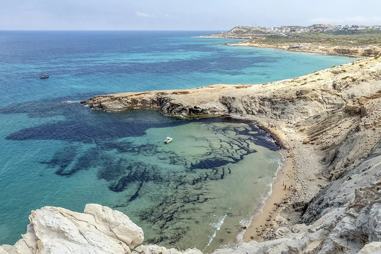 Enseada de água turquesa e verde entre costas de rocha clara, com um barco pequeno, uma faixa de praia com pessoas e casas no alto ao fundo, no Cap Blanc, perto de Bizerta.