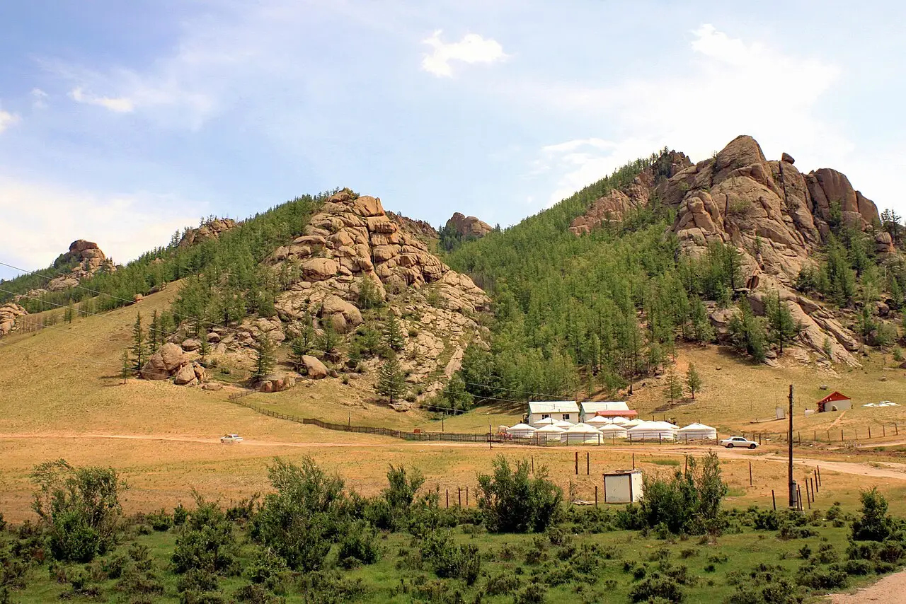 Colinas de rocha e floresta de coníferas com um acampamento de tendas brancas redondas no vale, no Parque Nacional Gorkhi-Terelj, com estrada de terra e carros pequenos.
