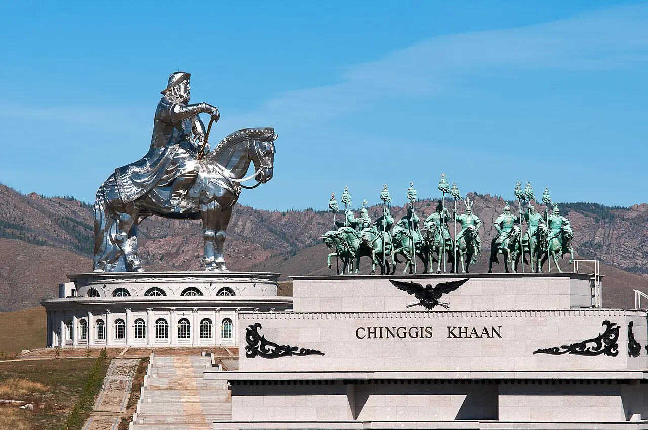 A estátua equestre de Chinggis Khaan, em aço polido, sobre um edifício circular com colunas, e um grupo de cavaleiros em bronze sobre a base com o nome, diante de colinas.