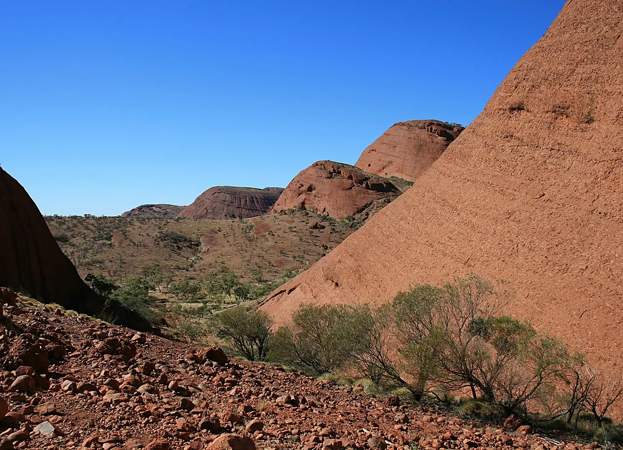 Vale entre domos de pedra vermelha em Kata Tjuta, com cascalho no chão e árvores baixas, sem pessoas.