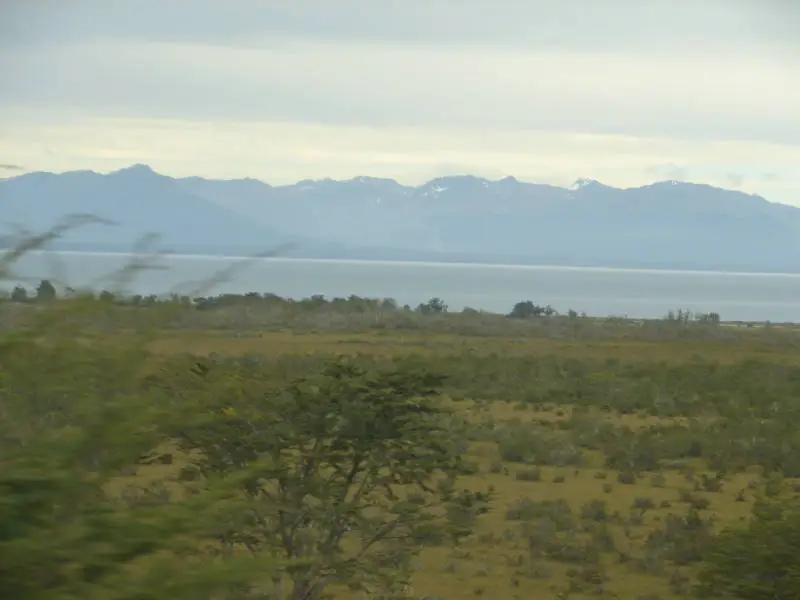 Campo com arbustos, uma faixa de água e montanhas com neve nos cumes.