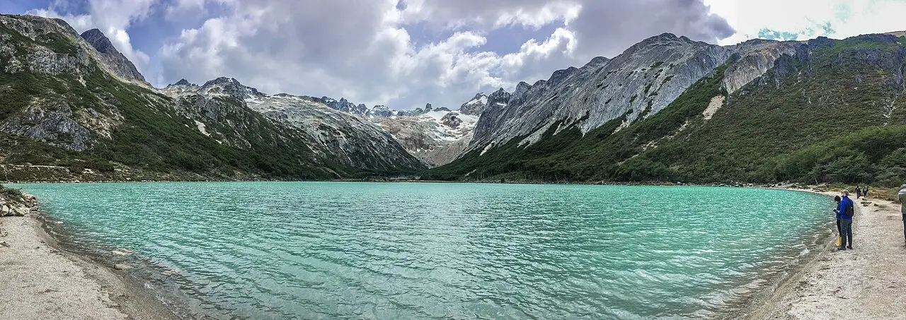 Panorama da Laguna Esmeralda, de água verde-turquesa, cercada por montanhas de rocha cinza com neve nos picos, e duas pessoas na margem à direita.