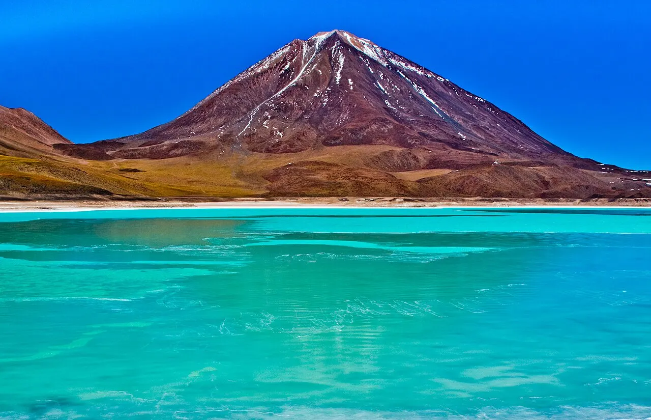 Água turquesa da Laguna Verde, com uma faixa clara de sal e um cone marrom e branco atrás.