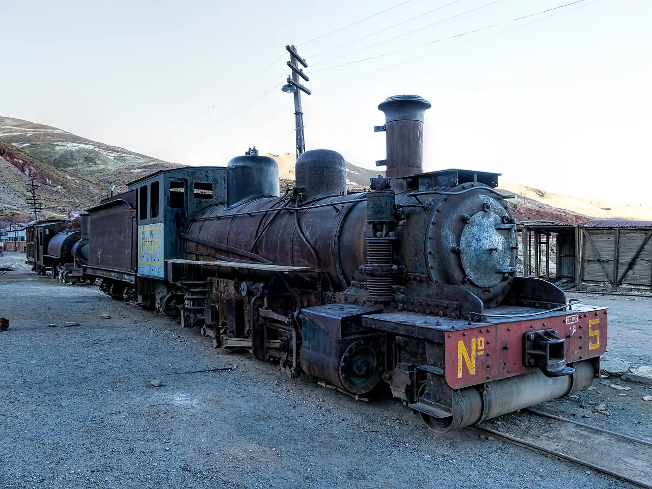 Locomotiva a vapor antiga e enferrujada, com o número 5 na frente, estacionada num pátio de cascalho, com vagões de madeira e morros secos ao fundo, em Pulacayo.