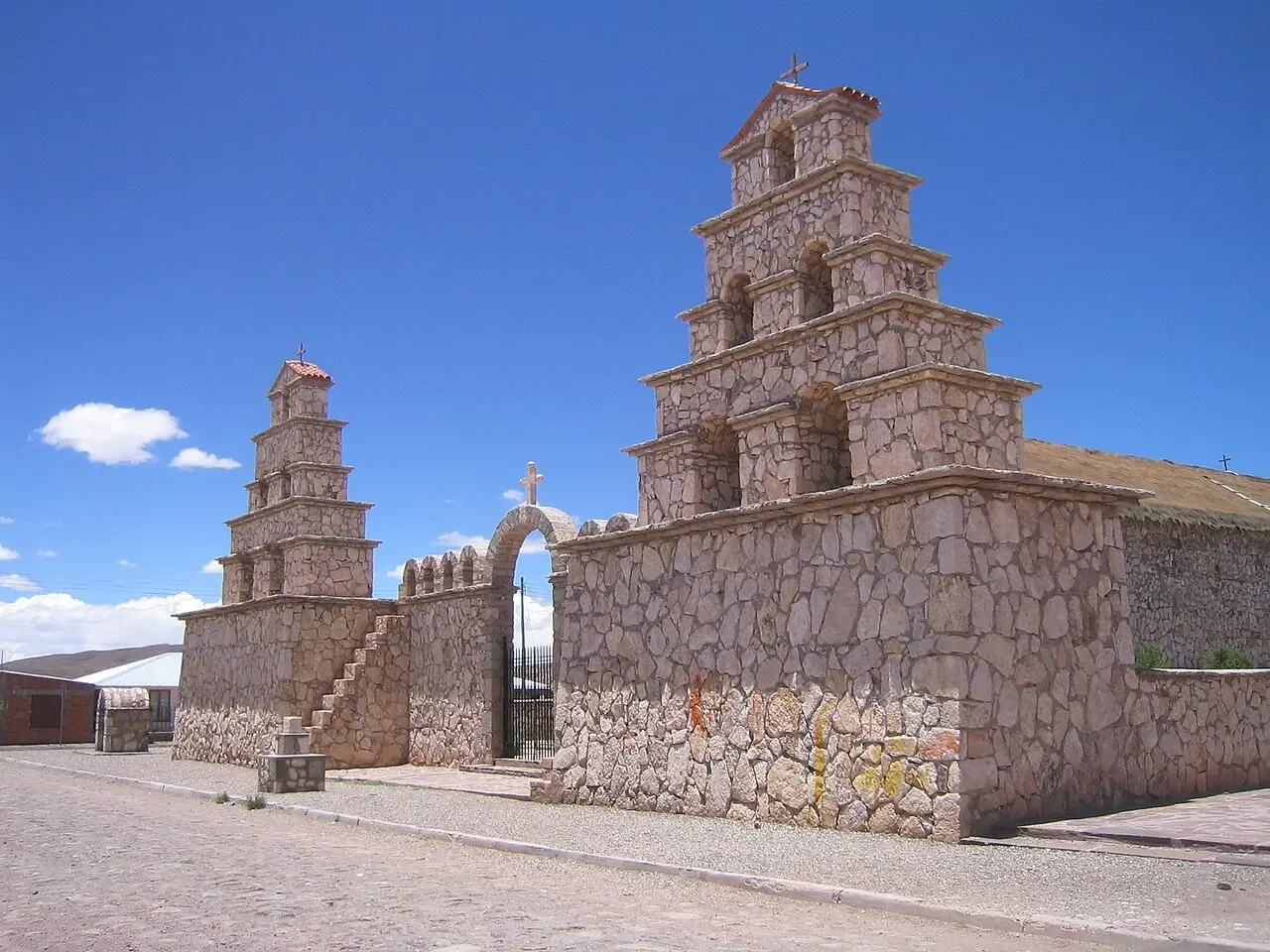Igreja de pedra com dois campanários escalonados e um portão em arco, numa rua de paralelepípedos, sob céu azul intenso, em San Cristóbal.