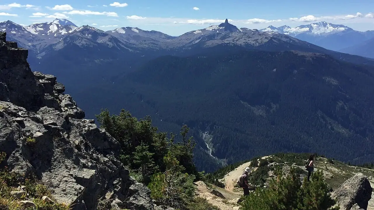 Vista de uma crista rochosa sobre um vale de floresta densa, com picos com neve e uma ponta escura ao fundo e duas pessoas caminhando por uma trilha, em Whistler.