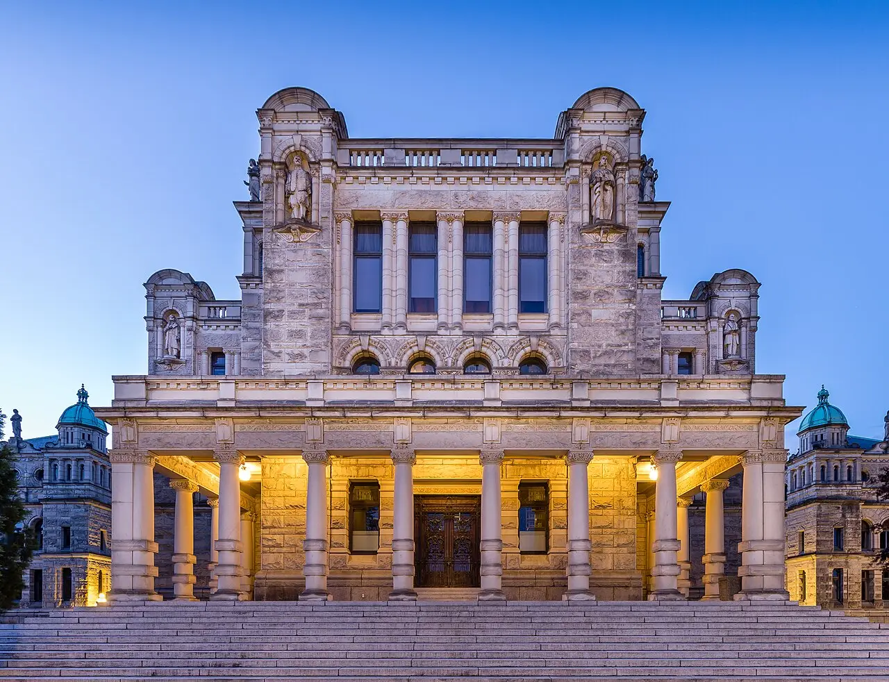 Fachada de pedra clara do Parlamento da Colúmbia Britânica ao entardecer, com colunas iluminadas na entrada, estátuas em nichos e uma escadaria larga na frente, em Victoria.