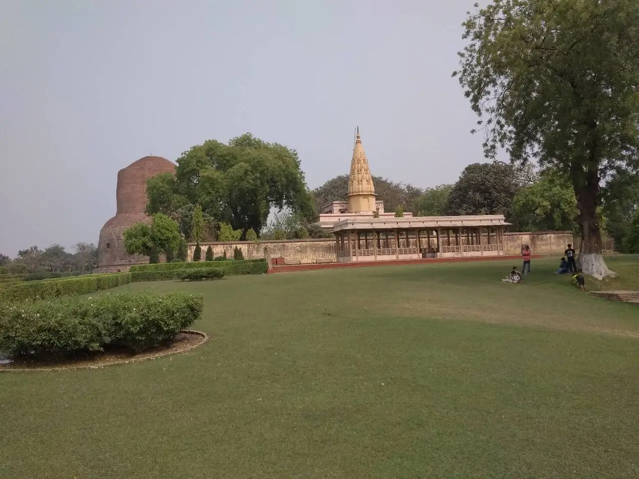 Gramado amplo com um estupa de tijolo à esquerda ao fundo e um pavilhão de pedra clara com torre pontuda, e algumas pessoas sentadas à sombra de uma árvore, em Sarnath.
