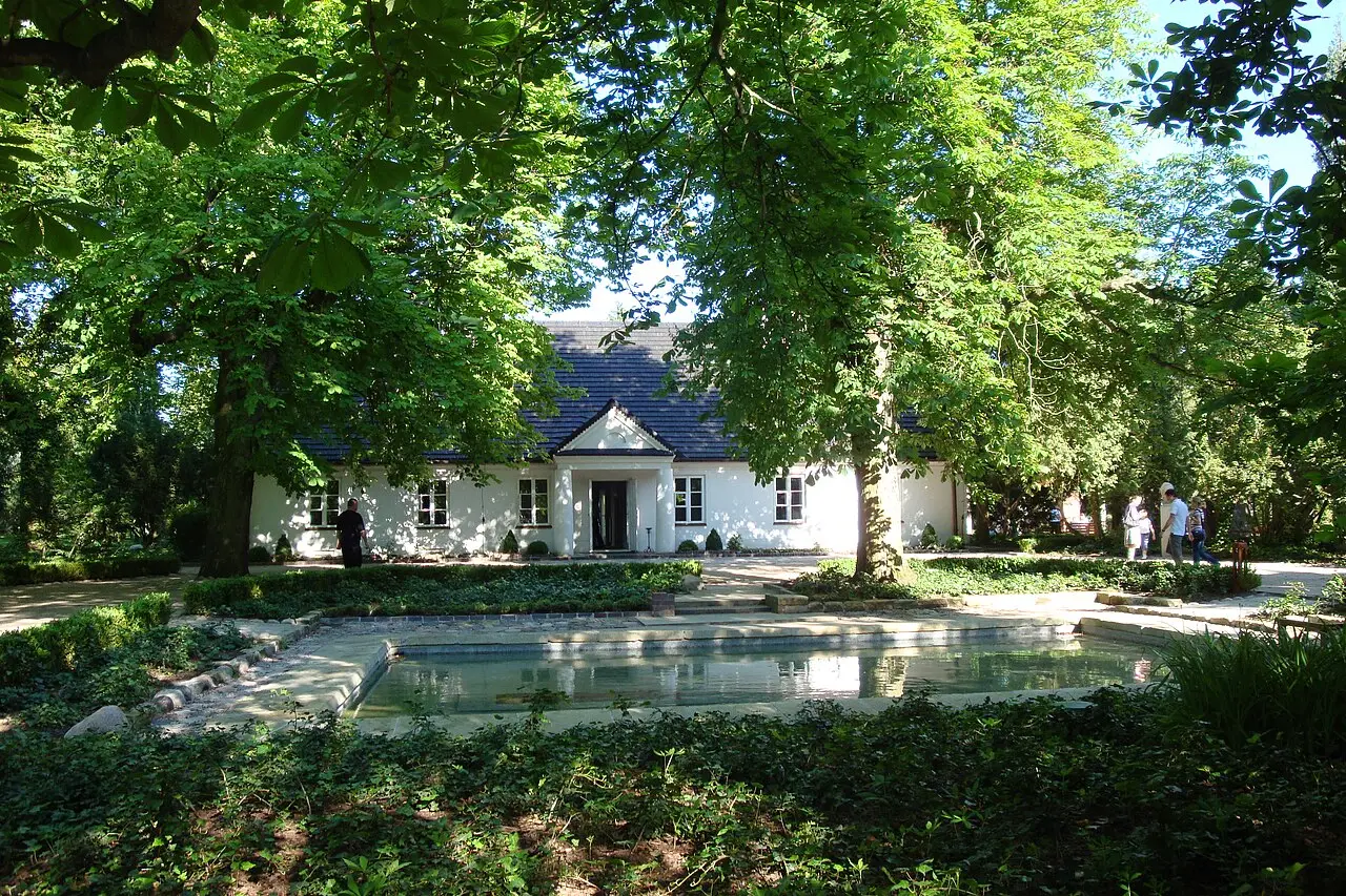Casa branca de um só andar com telhado escuro e pórtico de colunas, cercada de árvores grandes e de um tanque raso, com visitantes caminhando pelo parque, em Żelazowa Wola.