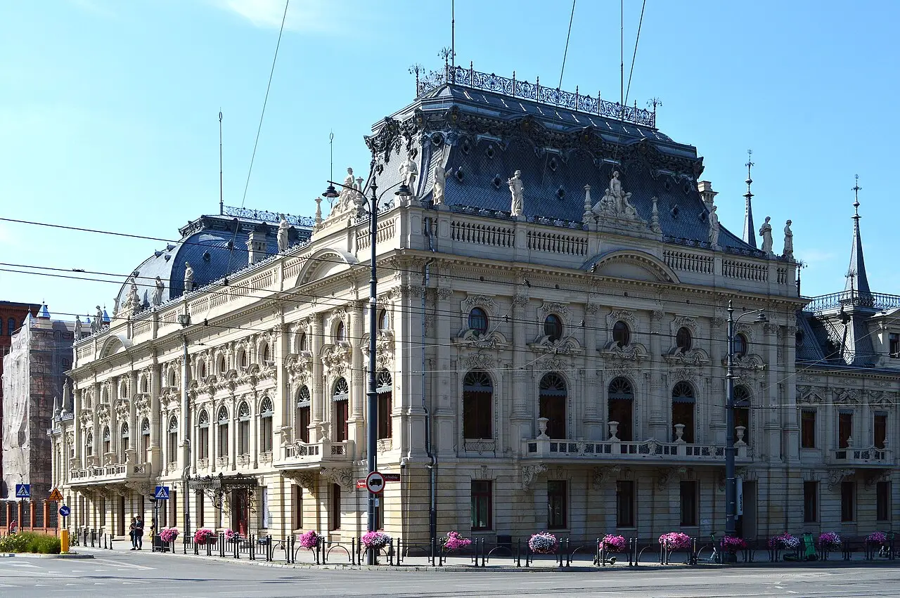 Esquina do Palácio Poznański em Łódź, edifício de fachada clara com estátuas na balaustrada e telhados de ardósia azulada, sobre uma rua com canteiros de flores.