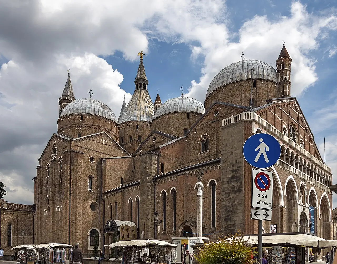 A Basílica de Santo Antônio de Pádua, de tijolo, com várias cúpulas cinzentas e torres pontudas, e barracas de lembranças e placas de trânsito em primeiro plano.