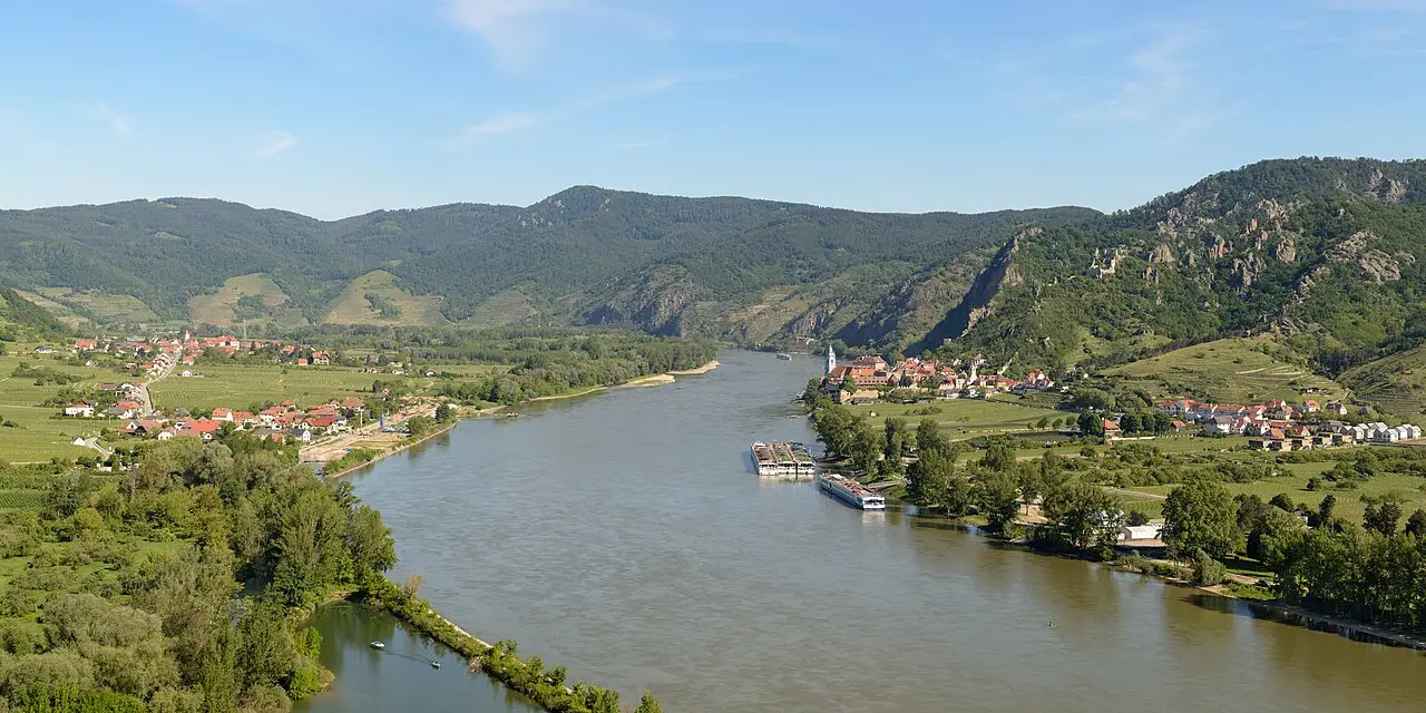O Danúbio numa curva larga entre colinas cobertas de floresta e vinhedos, com o povoado de Dürnstein na margem direita e dois barcos de cruzeiro atracados, na Wachau.