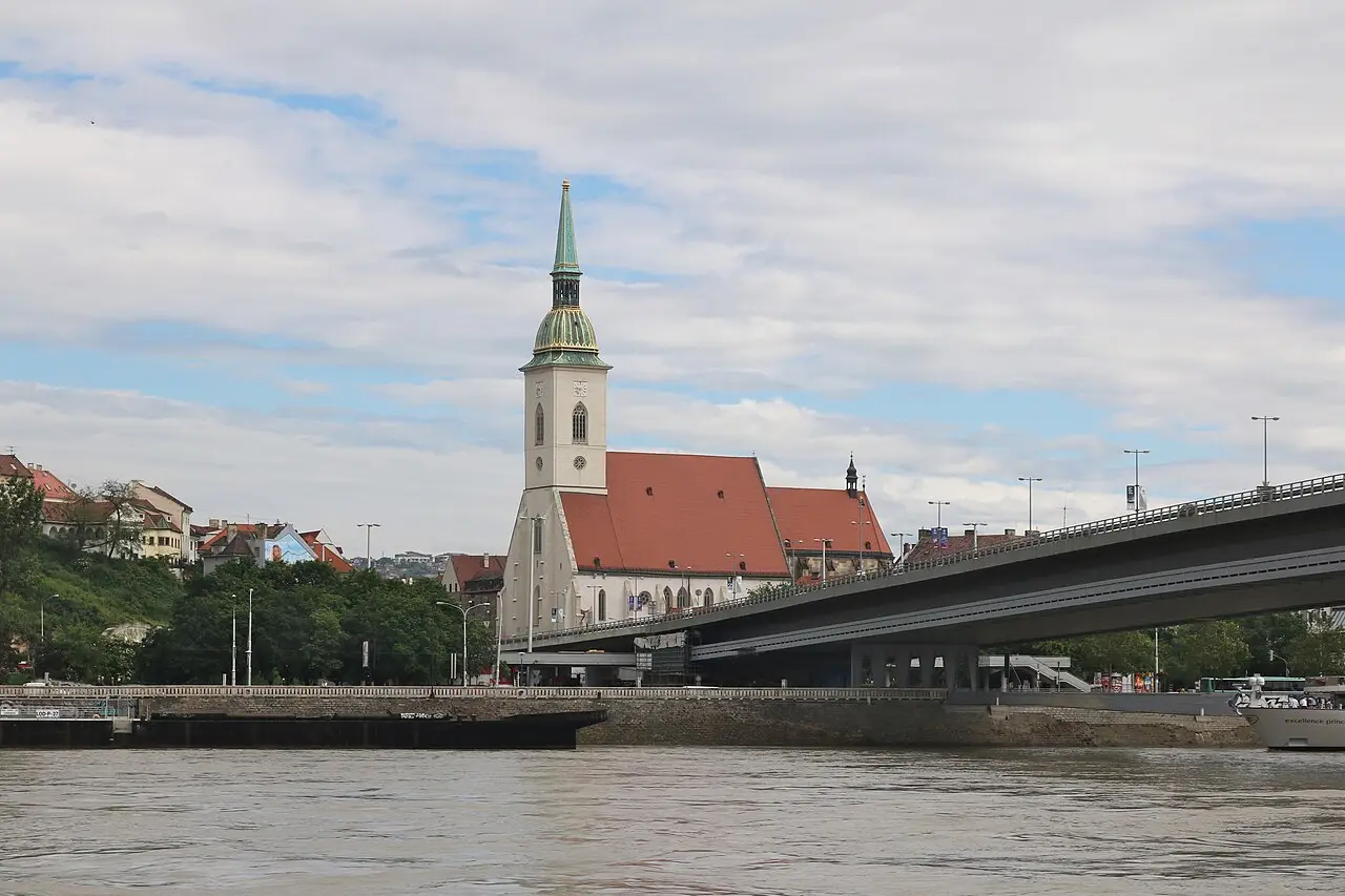 St Martin’s Cathedral in Bratislava, with a tall tower with a green dome and a red roof, beside an elevated road bridge, seen from the Danube.
