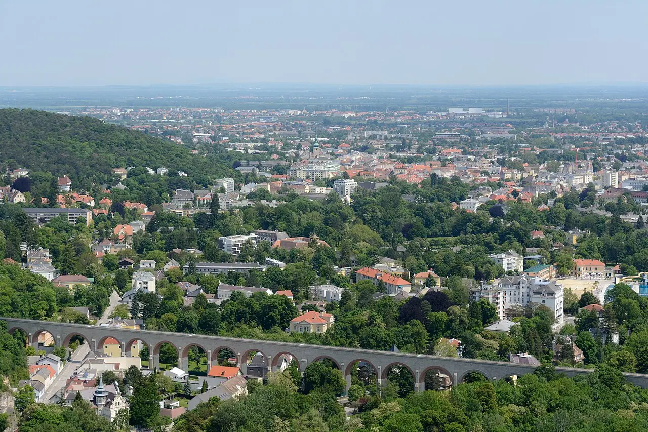 Vista aérea de Baden bei Wien, com casas de telhado laranja entre árvores, um aqueduto de arcos de pedra cruzando o bairro e a planície ao fundo.