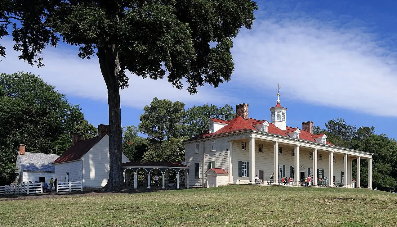 A casa principal de Mount Vernon, de tábuas claras e telhado vermelho com cúpula, com varanda de colunas onde há visitantes sentados, e um grande gramado à frente.