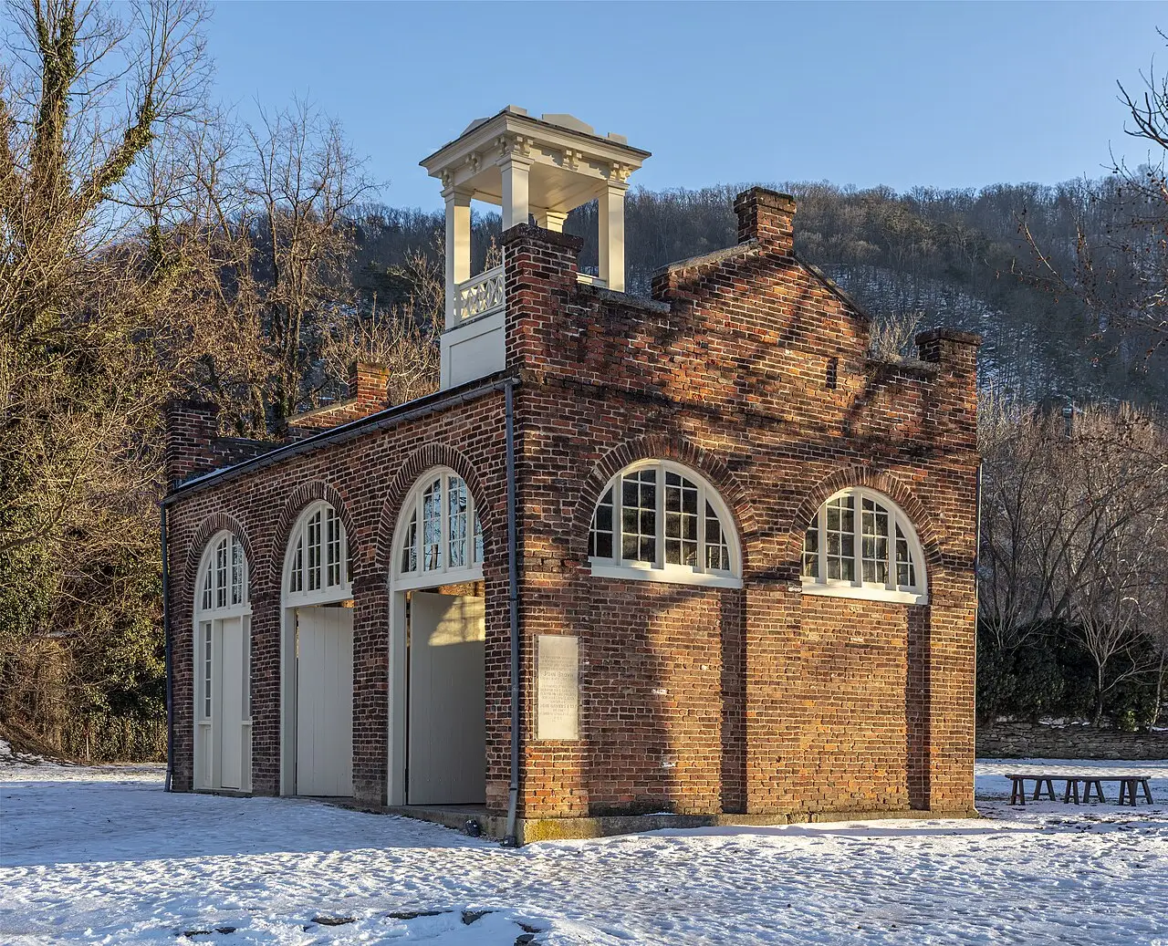 O edifício de tijolo conhecido como Fort de John Brown, com arcos brancos nas janelas e uma pequena torre no topo, sobre neve, com morros arborizados ao fundo, em Harpers Ferry.