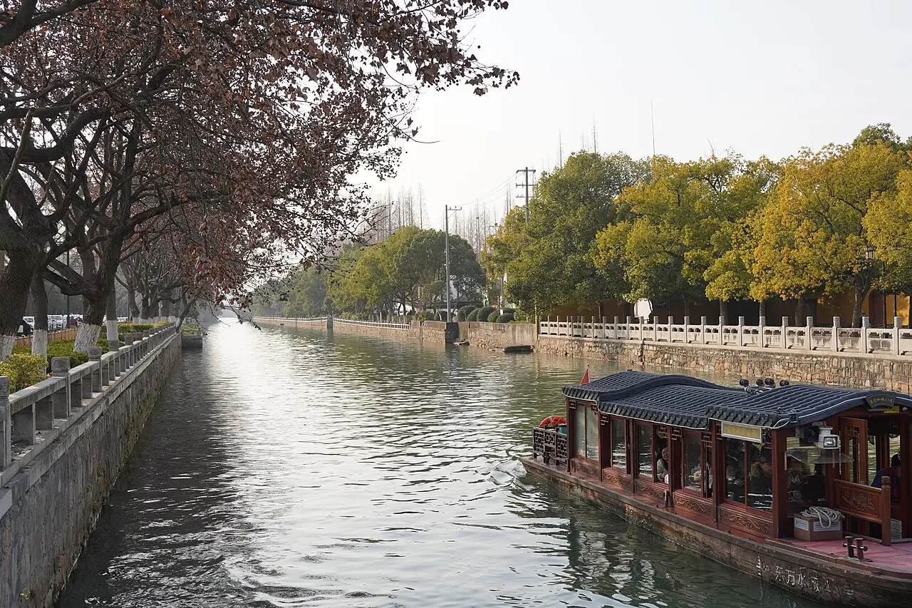 Canal de água calma em Suzhou, entre muretas de pedra e árvores de folhas amarelas, com um barco de turismo de telhado escuro à direita.