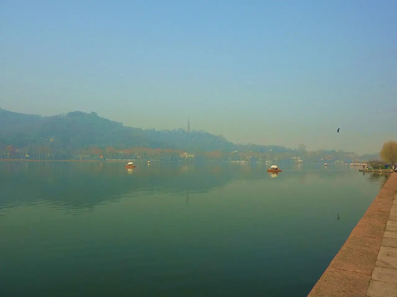 O lago Oeste, em Hangzhou, de água verde e calma sob uma névoa clara, com dois barcos pequenos, colinas ao fundo e uma pagoda esguia no alto, e um cais de pedra à direita.