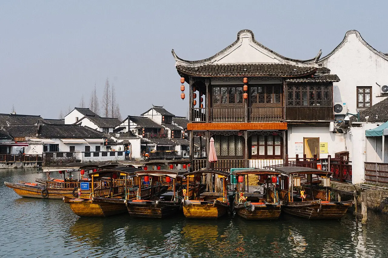 Barcos de madeira de proa curva atracados diante de casas tradicionais de dois andares, com telhado de telhas escuras, lanternas vermelhas e fachadas brancas, em Zhujiajiao.